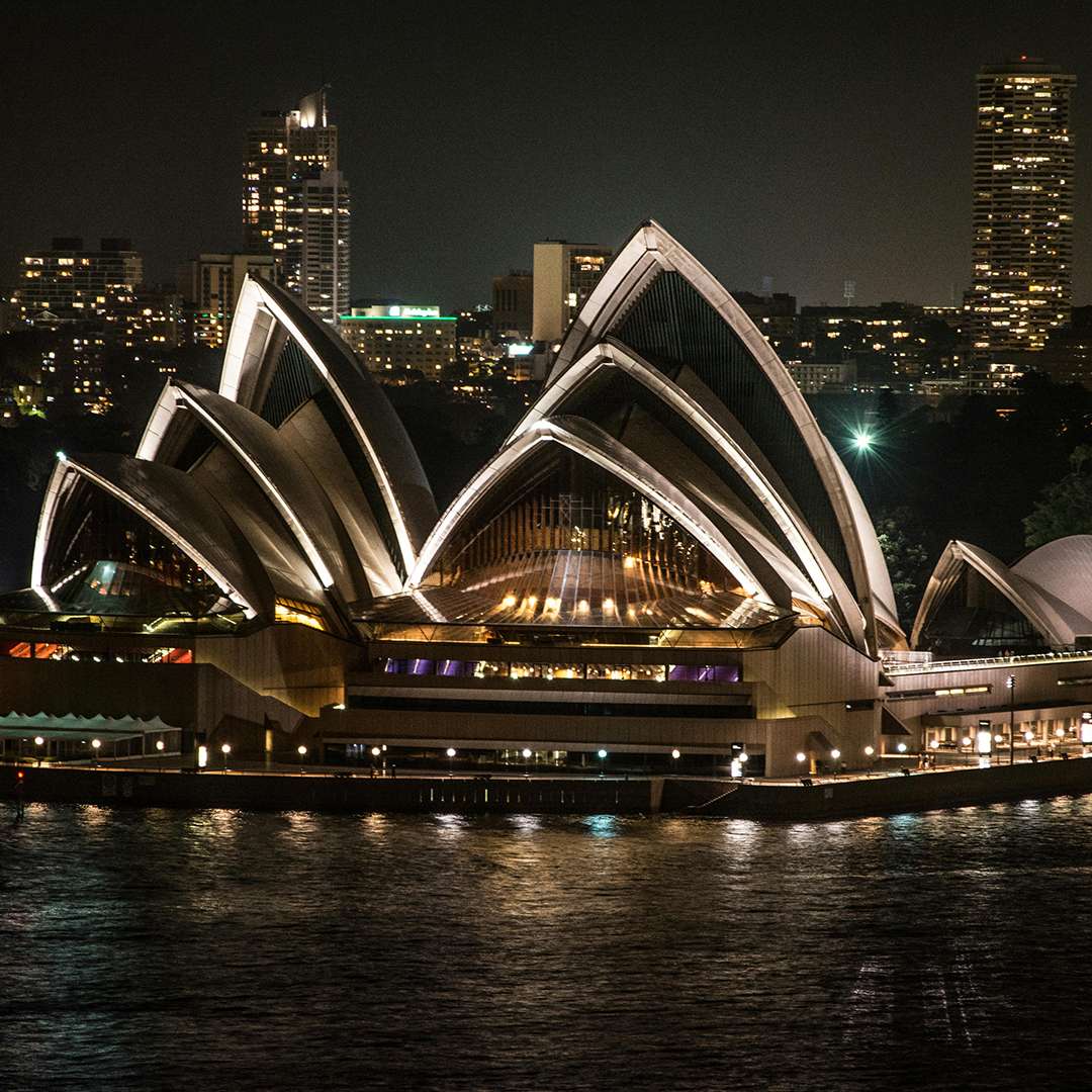 The Sydney Opera House at night, beautifully illuminated with lights against the city skyline. The shimmering reflection on the water showcases this iconic Australian landmark. ASK Consultants helps students study in Australia, making dreams come true.