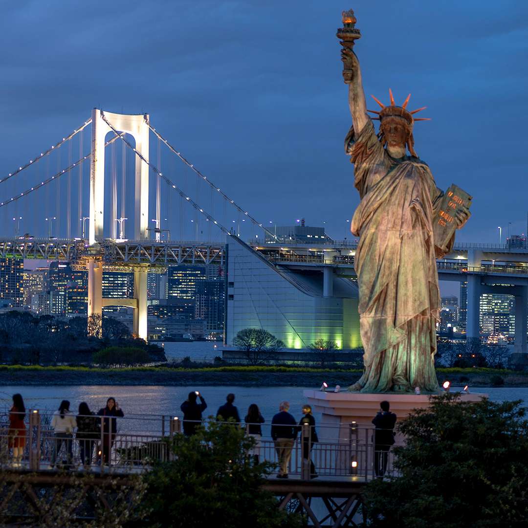 A nighttime view of a cityscape featuring a large, illuminated bridge with vibrant lights. In the foreground, there's a replica of the Statue of Liberty and people standing on a walkway, adding a sense of liveliness. The city skyline, with numerous lit buildings, serves as a striking backdrop. Used by ASK Consultants to highlight study abroad opportunities.